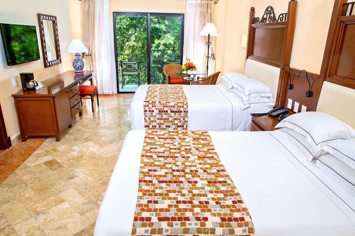 A hotel room with two beds, a desk with a lamp, a chair, a TV, and a balcony with chairs.