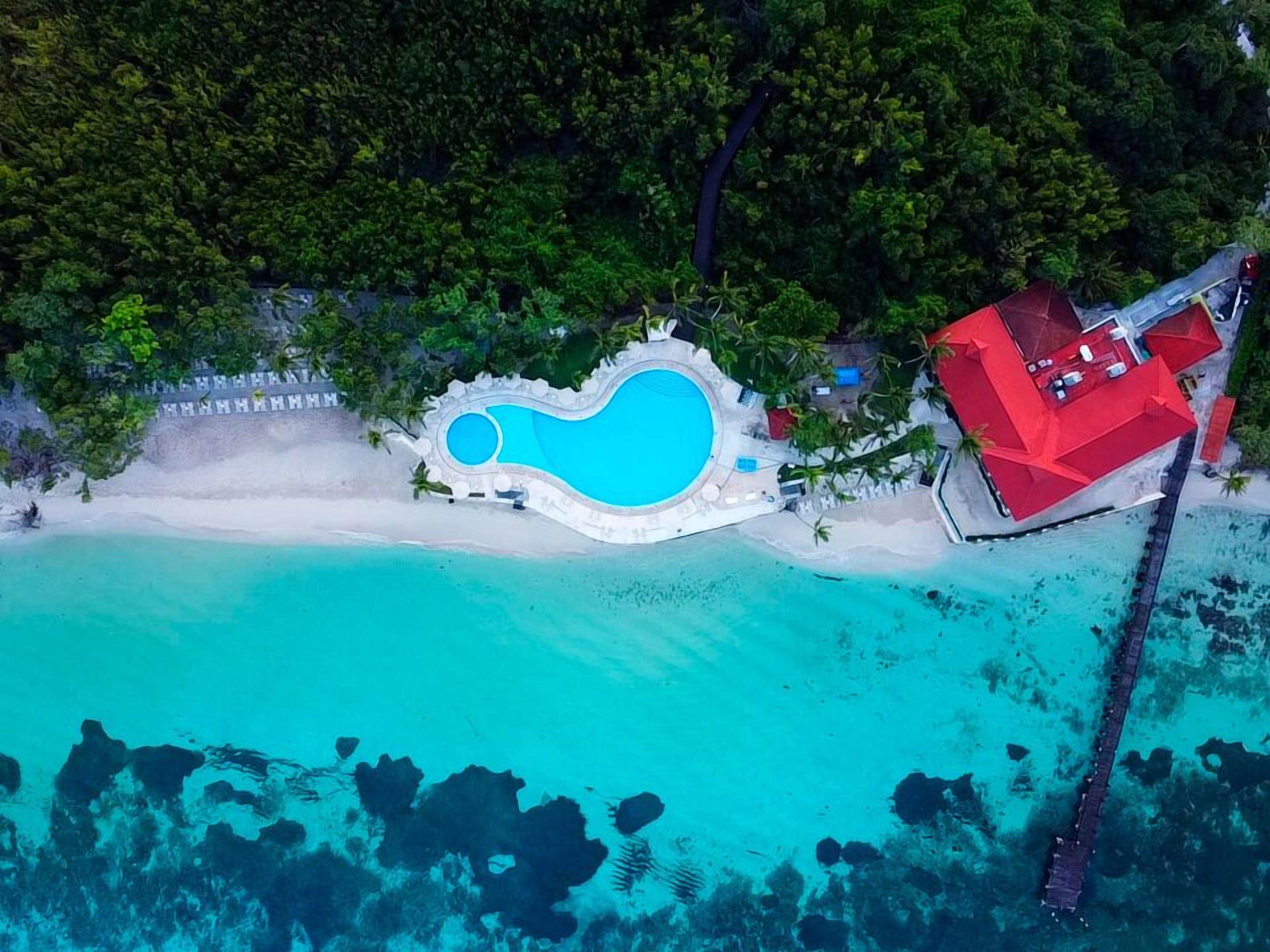 An aerial view of a resort with a large pool, a red building, and a beach.