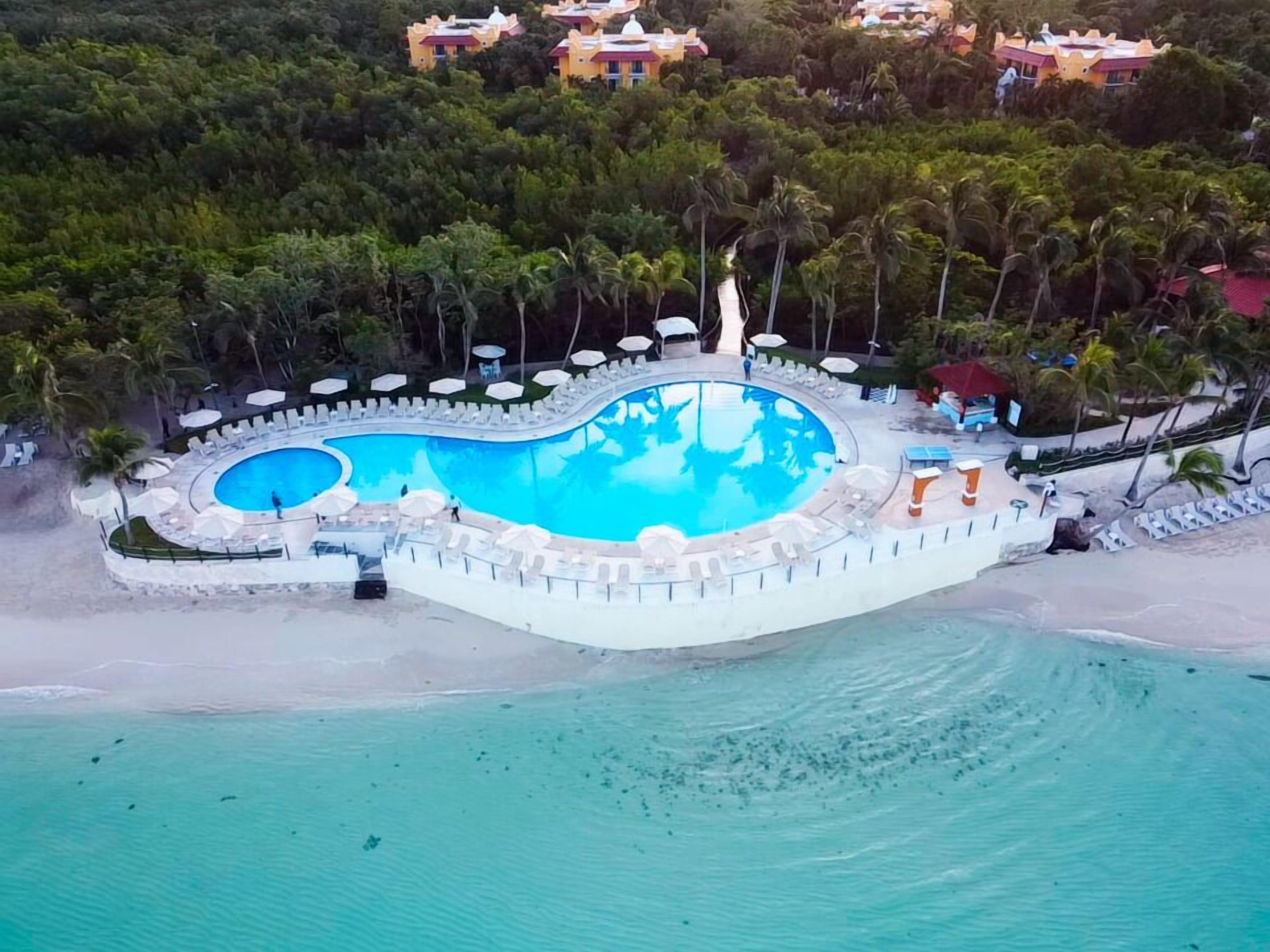 Vista aérea de un complejo turístico con una amplia piscina curva rodeada de sillones de playa y sombrillas, con palmeras y una playa de fondo.