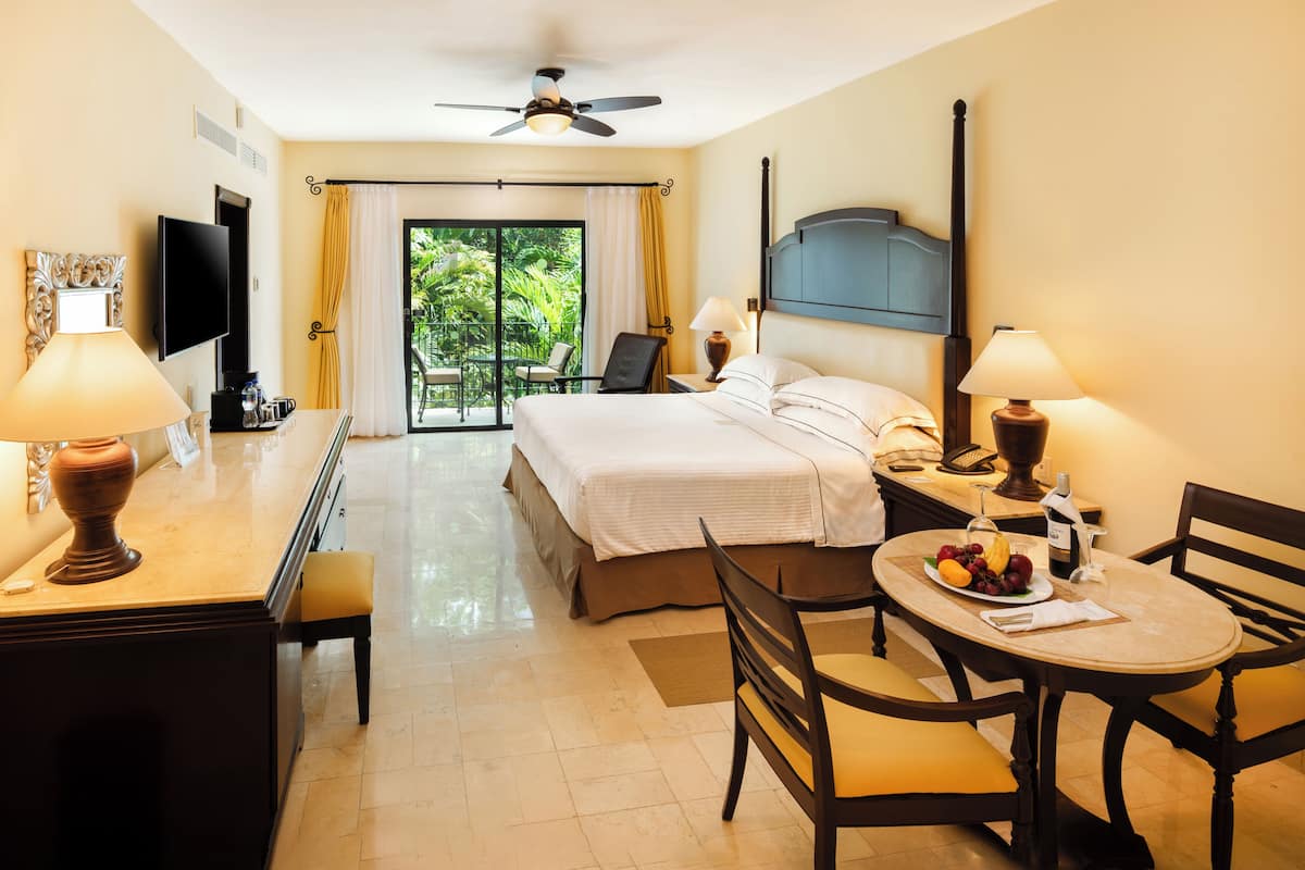 A hotel room with a bed, two bedside tables with lamps, a desk with a chair, a dining table with chairs, a television, and a balcony with curtains.