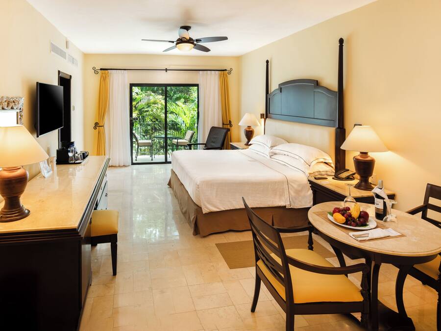 Quarto de hotel com cama, duas mesas de cabeceira com luminárias, escrivaninha com cadeira, mesa de jantar com cadeiras, televisão e varanda com cortinas.