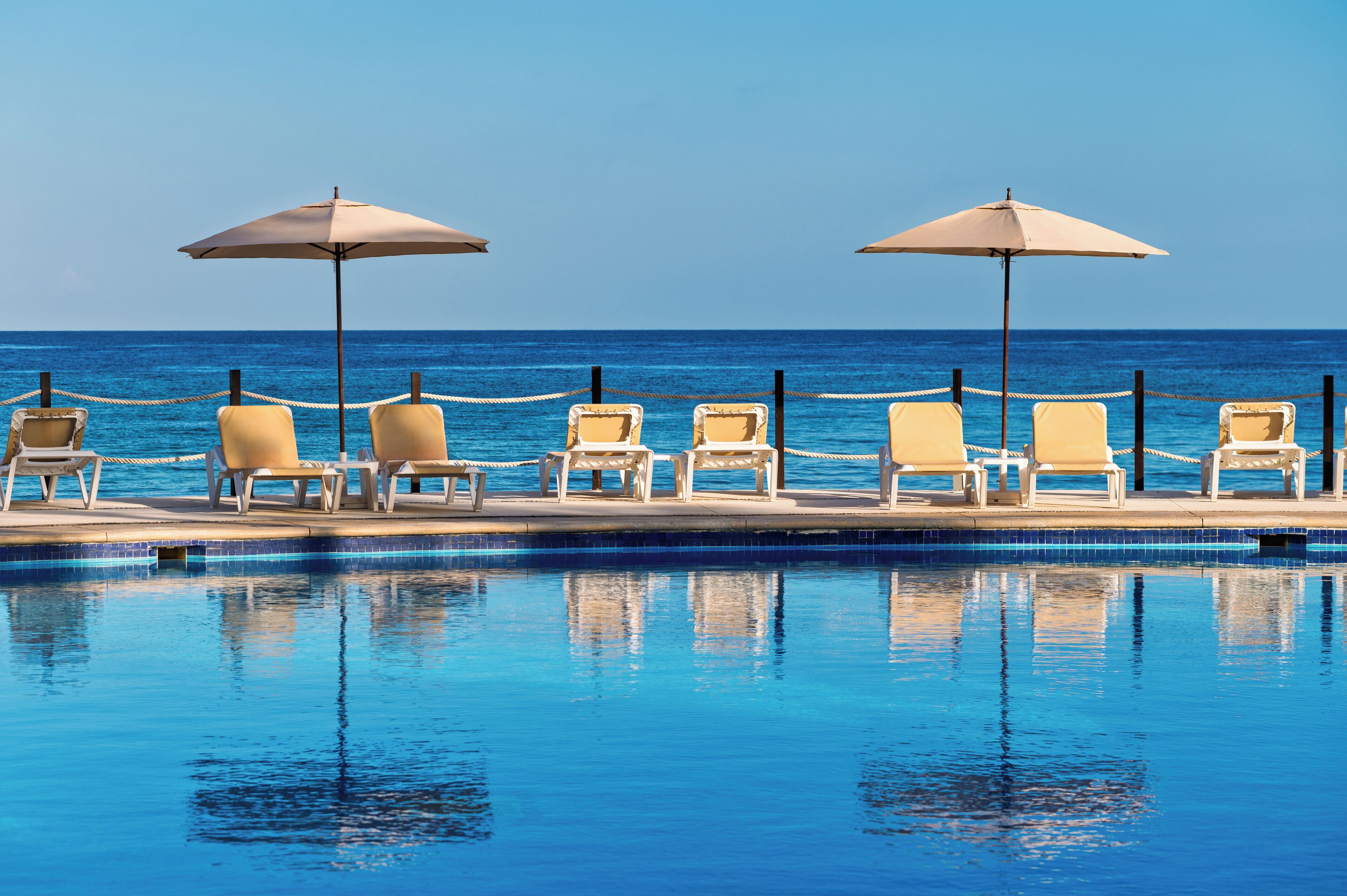 Zona junto a la piscina con sillones de descanso y sombrillas, una barrera de cuerda y vista al océano.