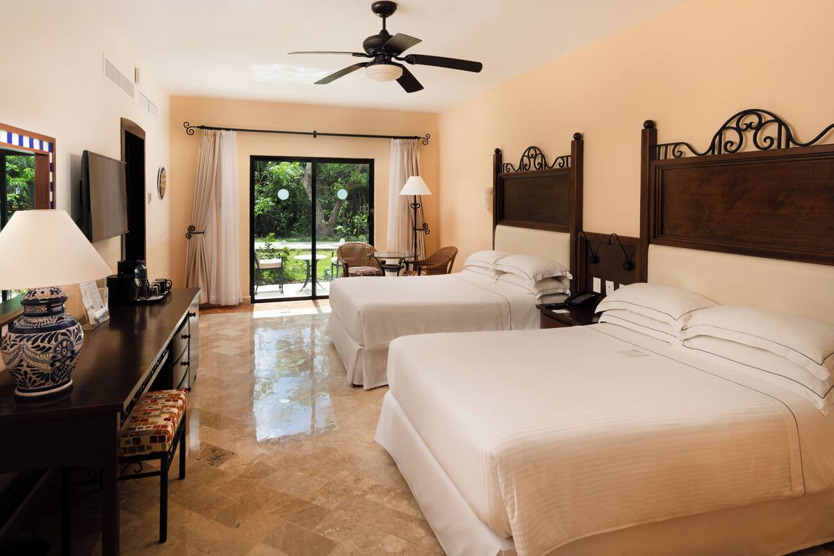 A hotel room with two beds, a ceiling fan, a TV, a desk with a lamp, and a view of the outdoors.