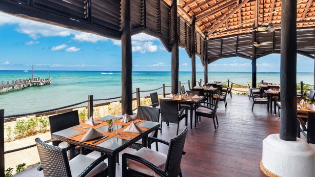 An outdoor dining area with tables and chairs, overlooking a clear blue sea and a pier. - Occidental Cozumel - All Inclusive (Cozumel)