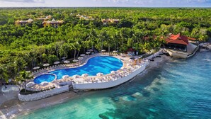 An aerial view of a resort with a large circular pool surrounded by lounge chairs and umbrellas, set against a backdrop of lush greenery and a clear blue sea. - Occidental Cozumel - All Inclusive (Cozumel)