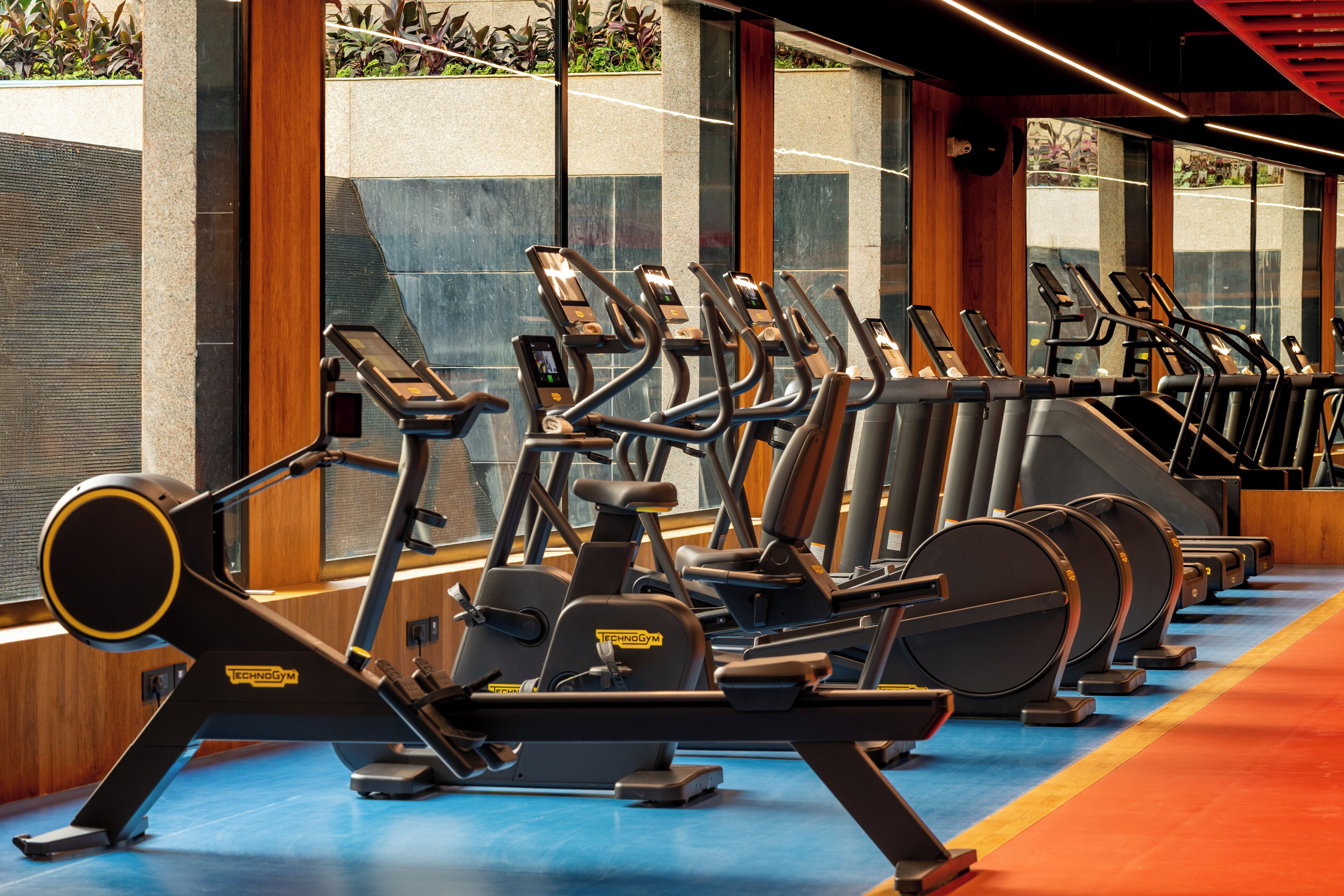 A row of stationary exercise bikes in a gym with large windows and a blue floor.