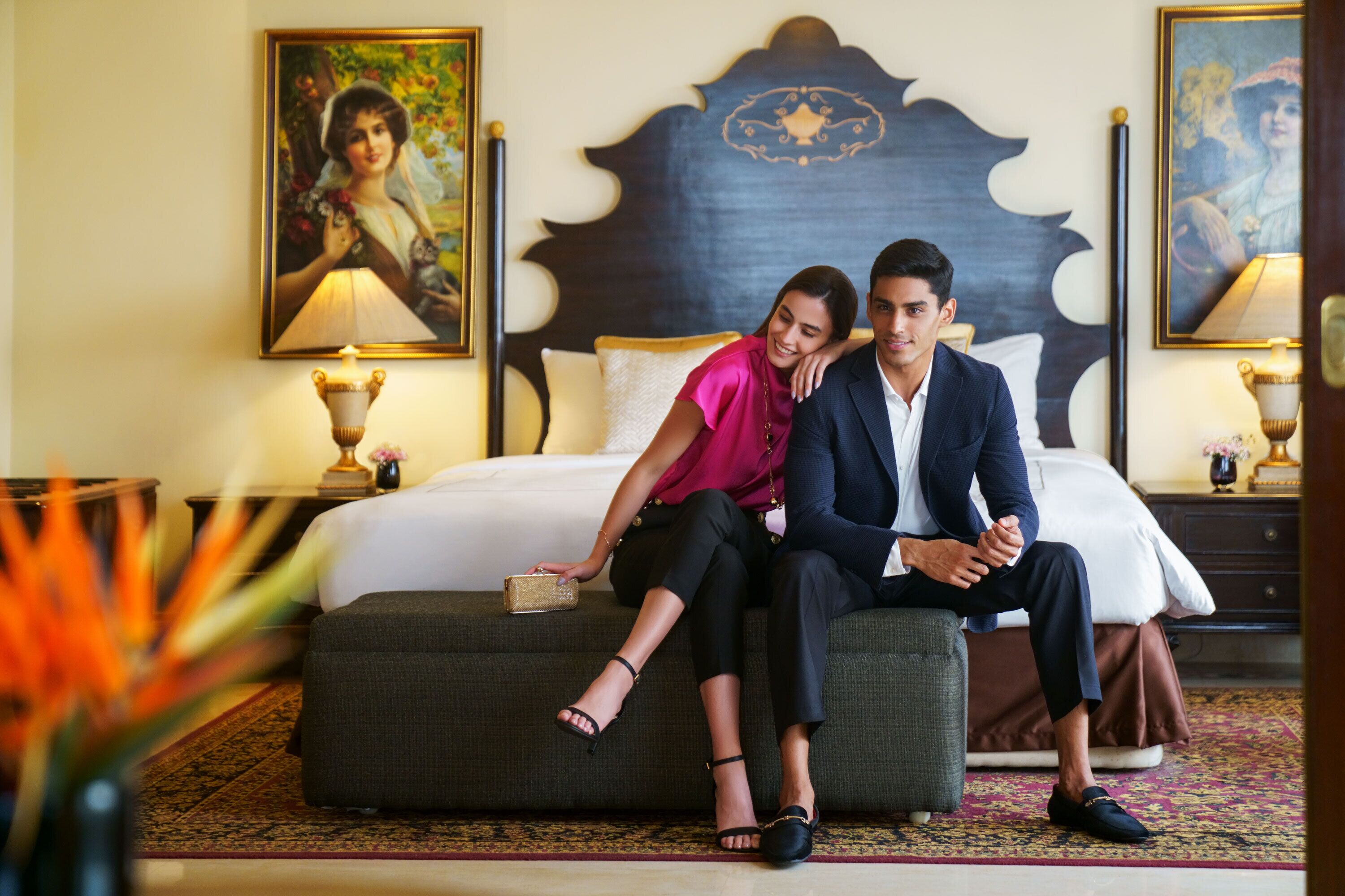 Un couple est assis sur un lit dans une chambre d’hôtel. On peut voir un grand et somptueux panneau de tête de lit, une table de chevet avec une lampe et un tableau accroché au mur.