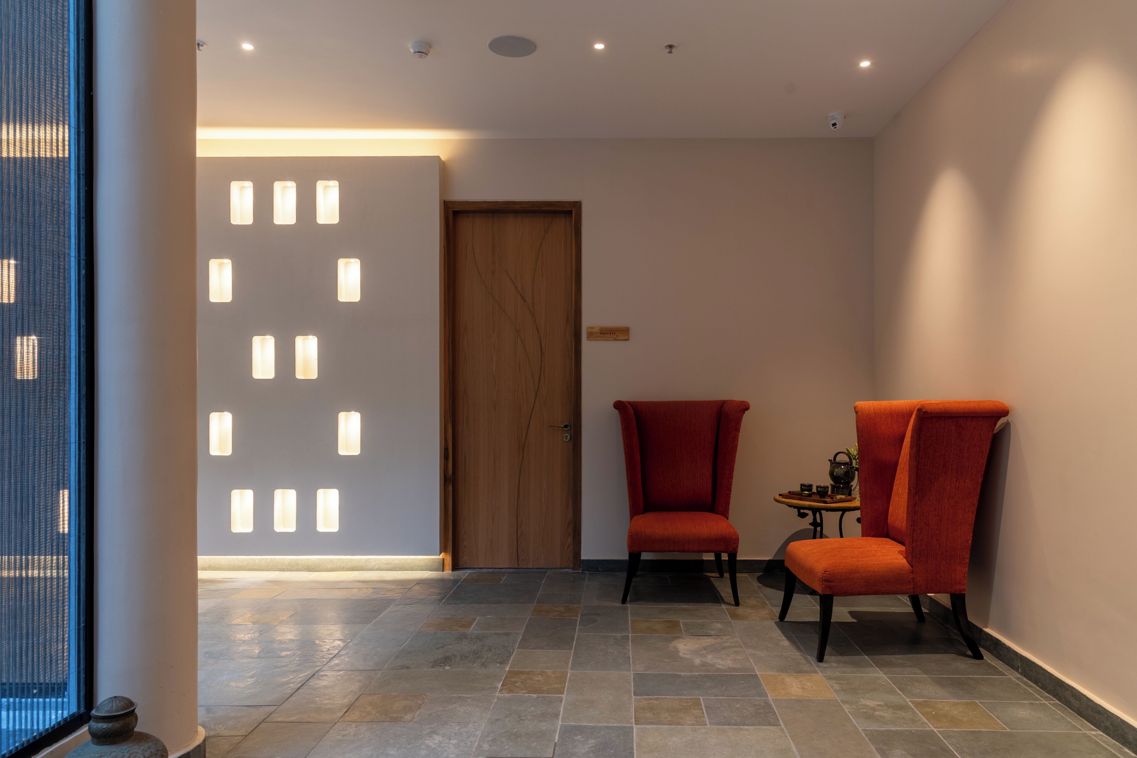 A hallway with a wooden door, two red chairs, a small table with a teapot, and a tiled floor.