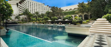 Una gran piscina con agua cristalina y azul, rodeada de sillas de descanso y sombrillas, con un edificio al fondo.