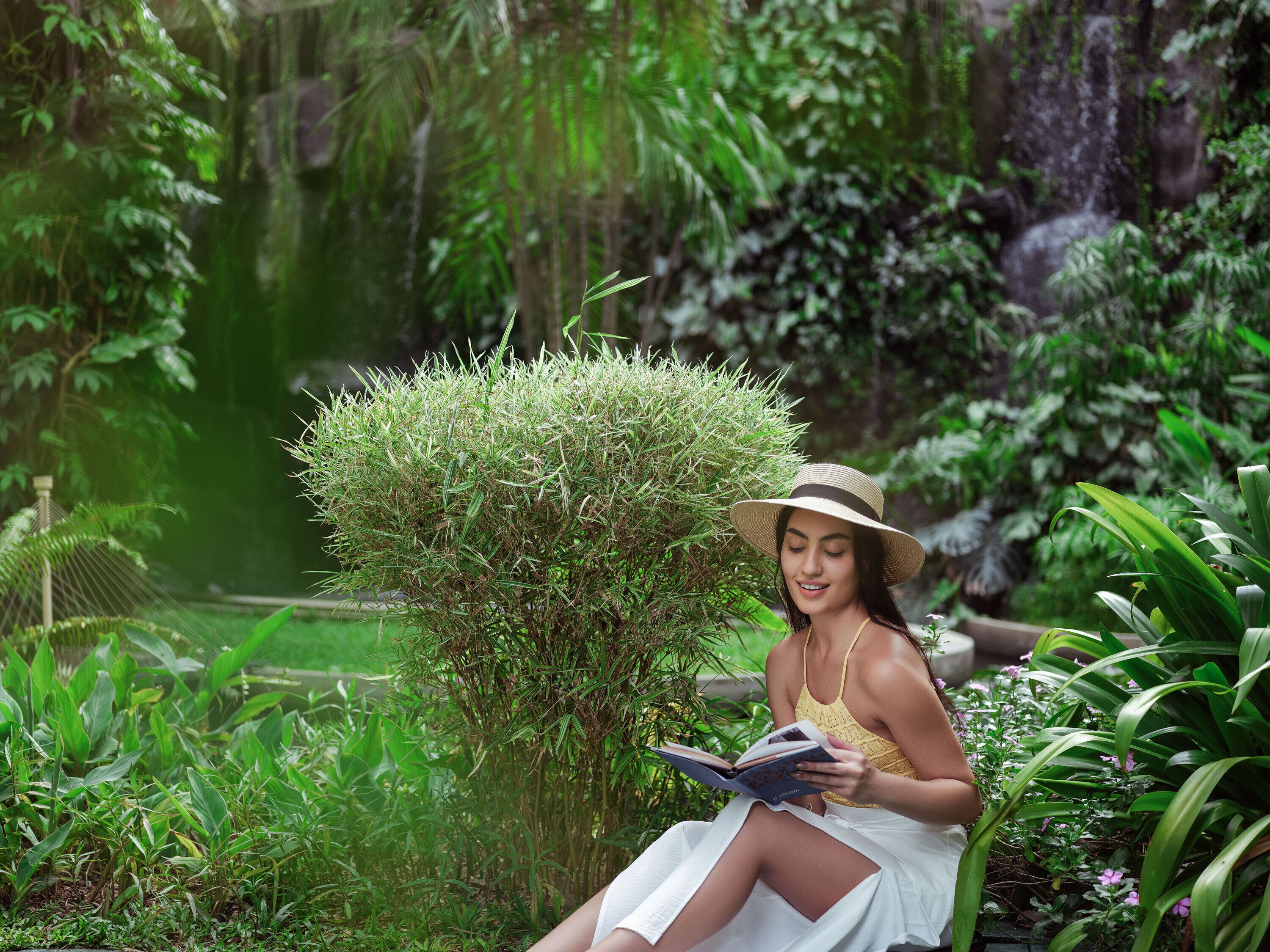 A woman reading a book in a lush garden with dense foliage and a small waterfall in the background.