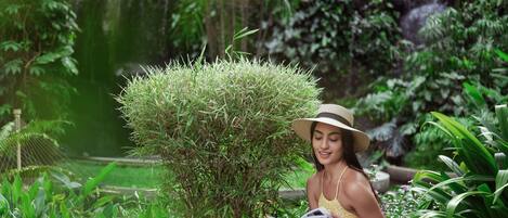 A woman reading a book in a lush garden with dense foliage and a small waterfall in the background.