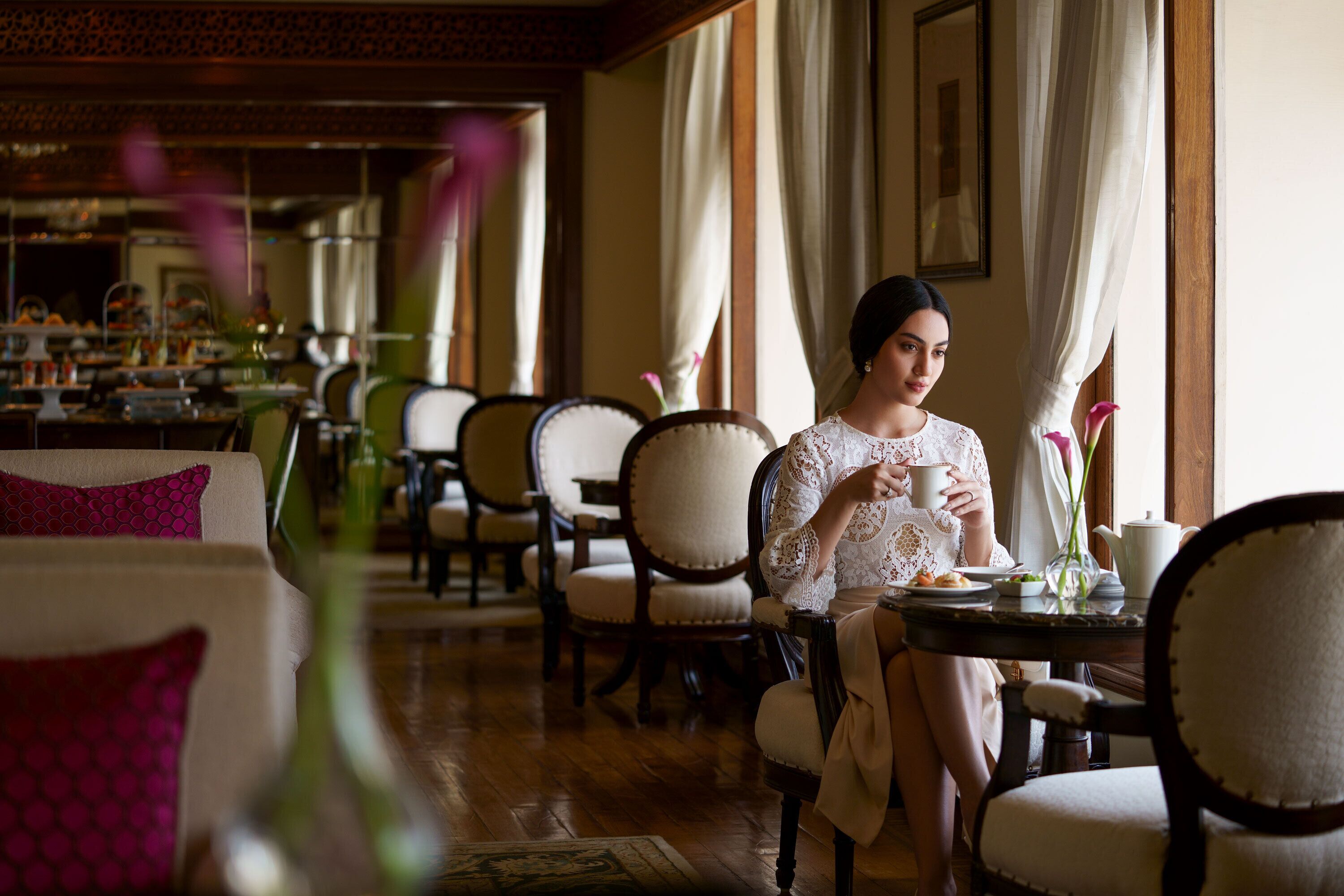 A woman seated at a table in a room with multiple chairs and a dining table, with a window and curtains in the background.