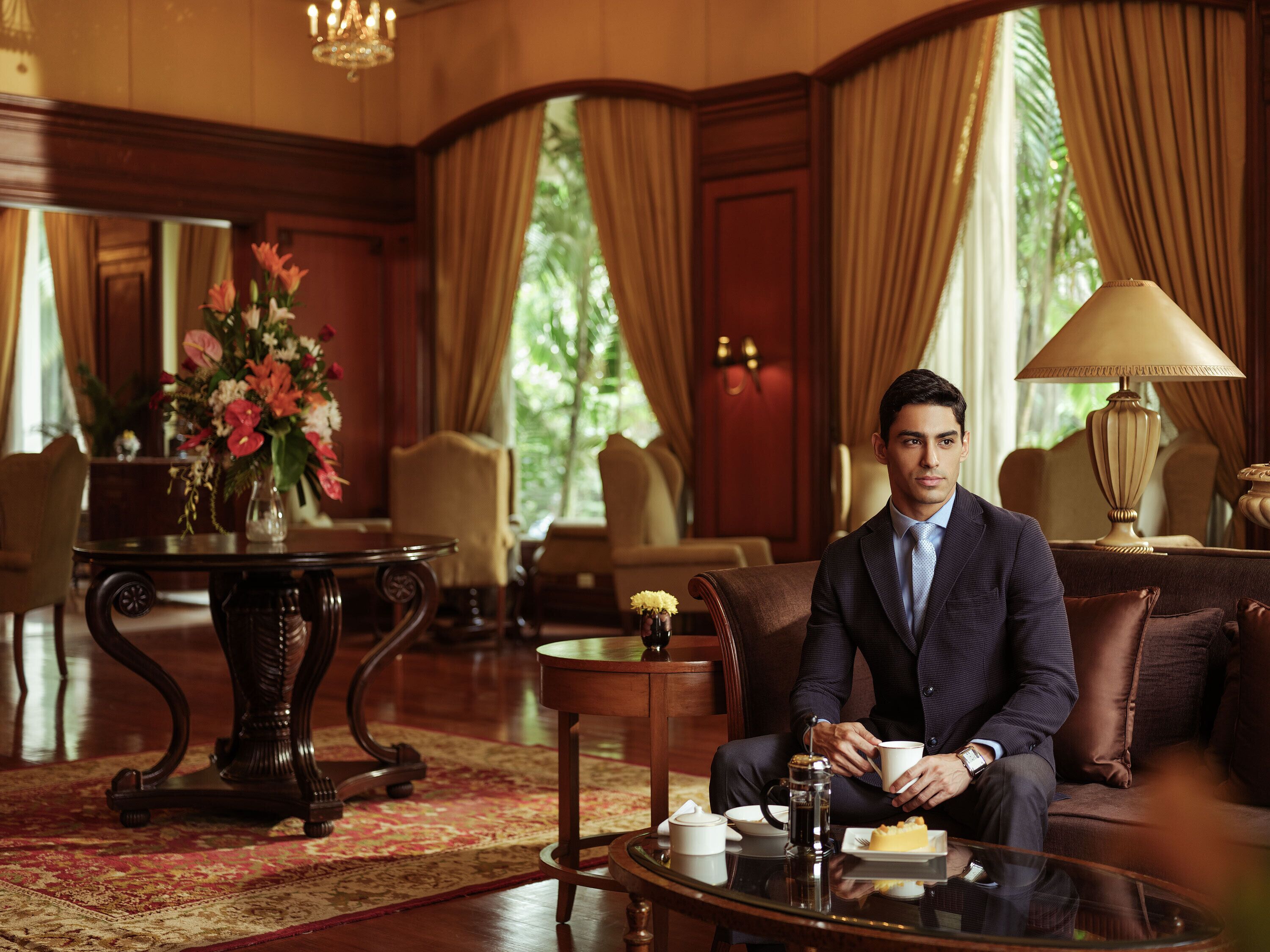 A man in a suit sitting on a couch in a well-furnished room with a coffee table, a vase of flowers, and a lamp.