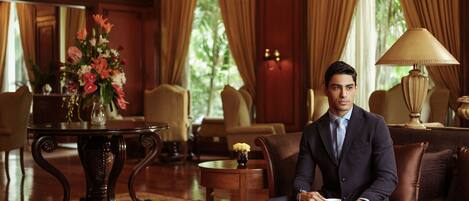 A man in a suit sitting on a couch in a well-furnished room with a coffee table, a vase of flowers, and a lamp.
