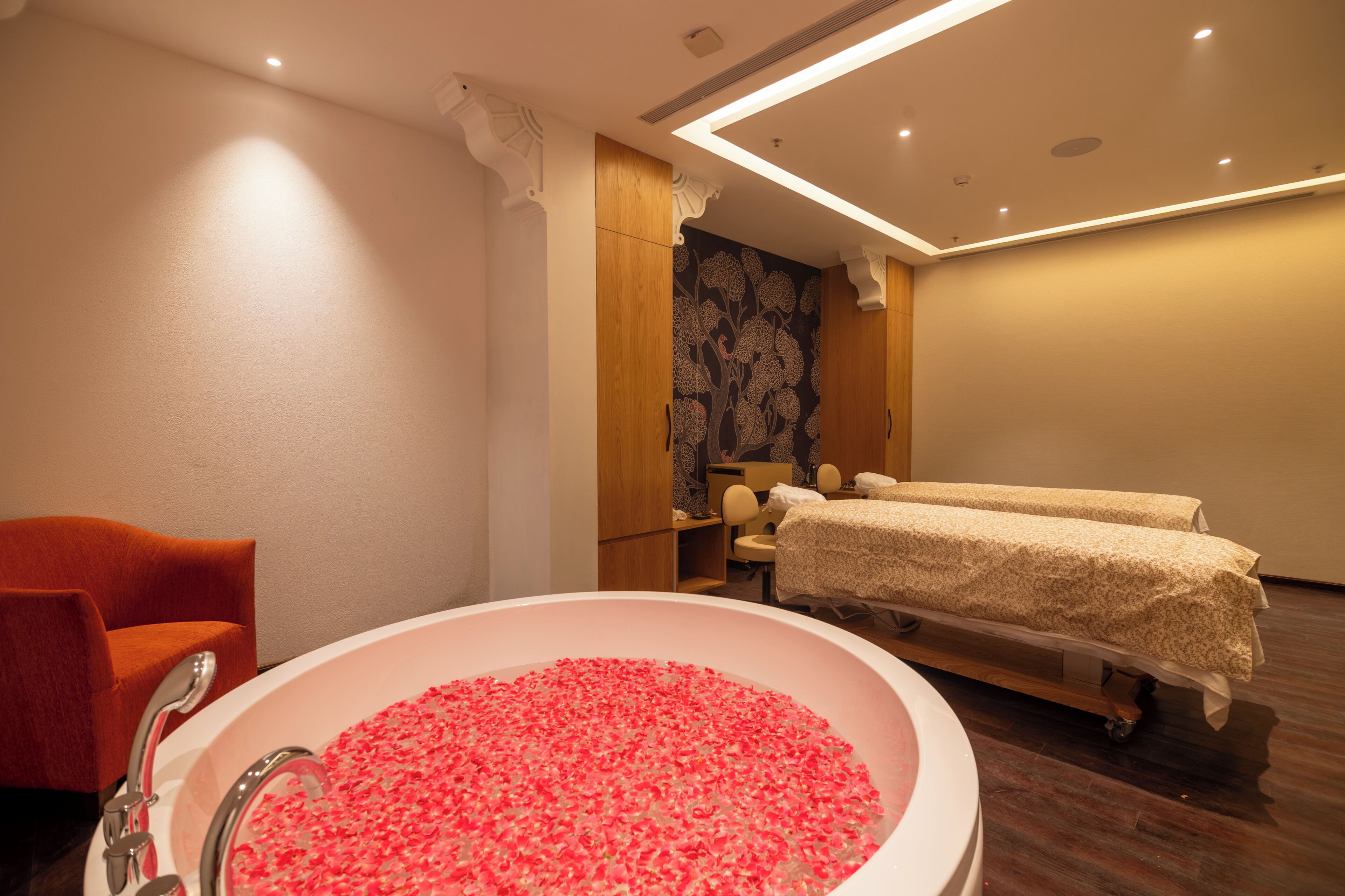 A room with a large bathtub filled with rose petals, a wooden cabinet, a massage table, and a red chair.
