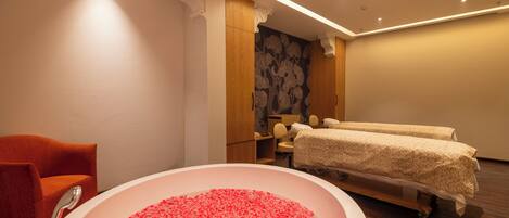 A room with a large bathtub filled with rose petals, a wooden cabinet, a massage table, and a red chair.