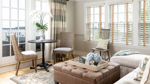 A cozy living room with a brown leather ottoman, wicker chairs, a round table, and a window with blinds. - Vineyard Square Hotel & Suites (Edgartown)