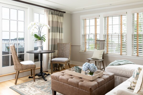 A cozy living room with a brown leather ottoman, wicker chairs, a round table, and a window with blinds.