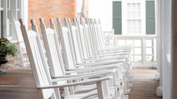 A row of white rocking chairs on a wooden deck with a brick wall and windows in the background.