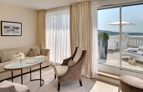 A modern living room with a sofa, armchair, and coffee table. A large window with curtains offers a view of the outdoors. - Grand Hôtel Spa & Thalasso à Saint-Jean-de-Luz (Saint-Jean-de-Luz)