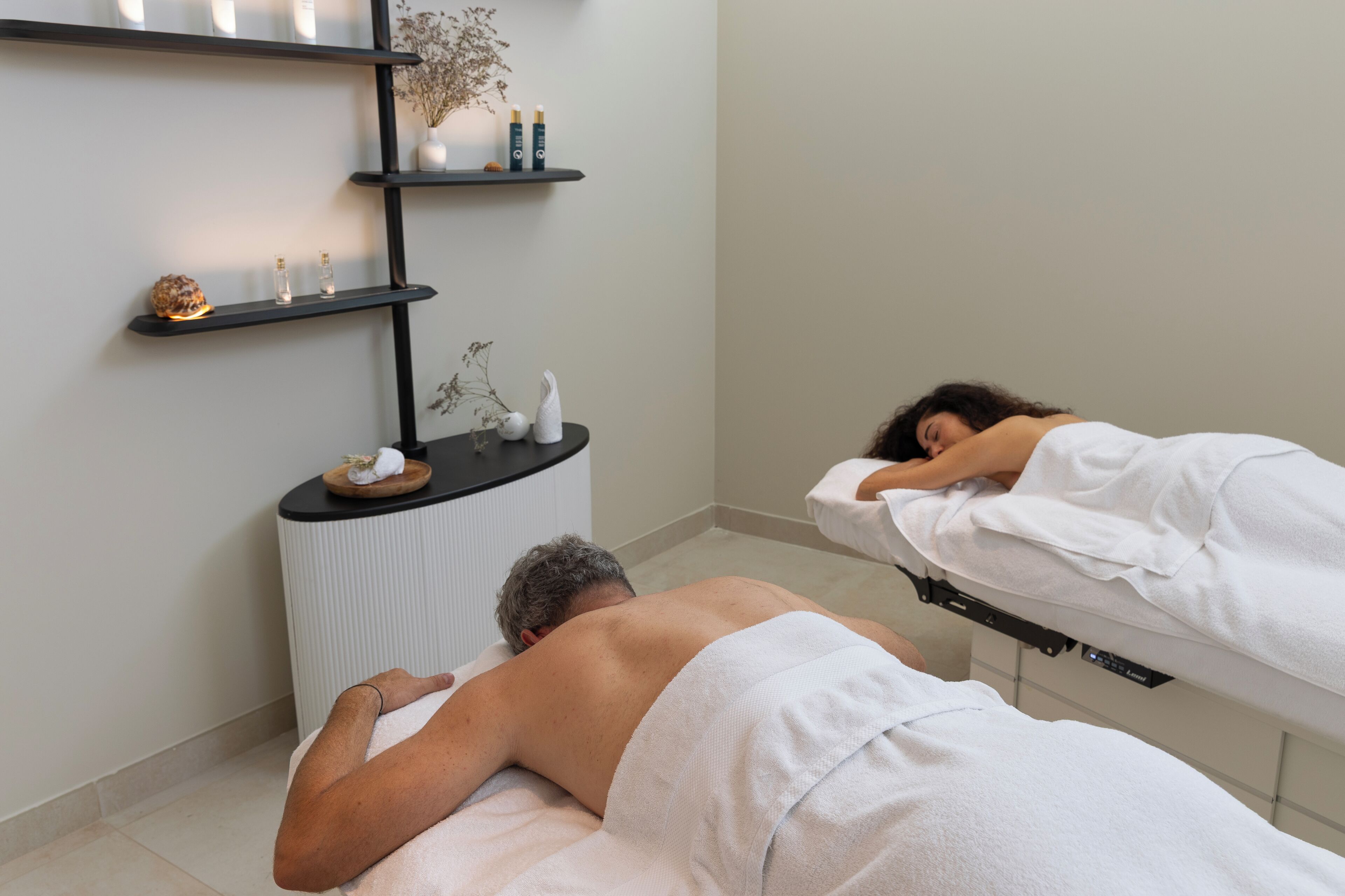 A spa room with two massage tables, a shelf with bottles, and a small plant.