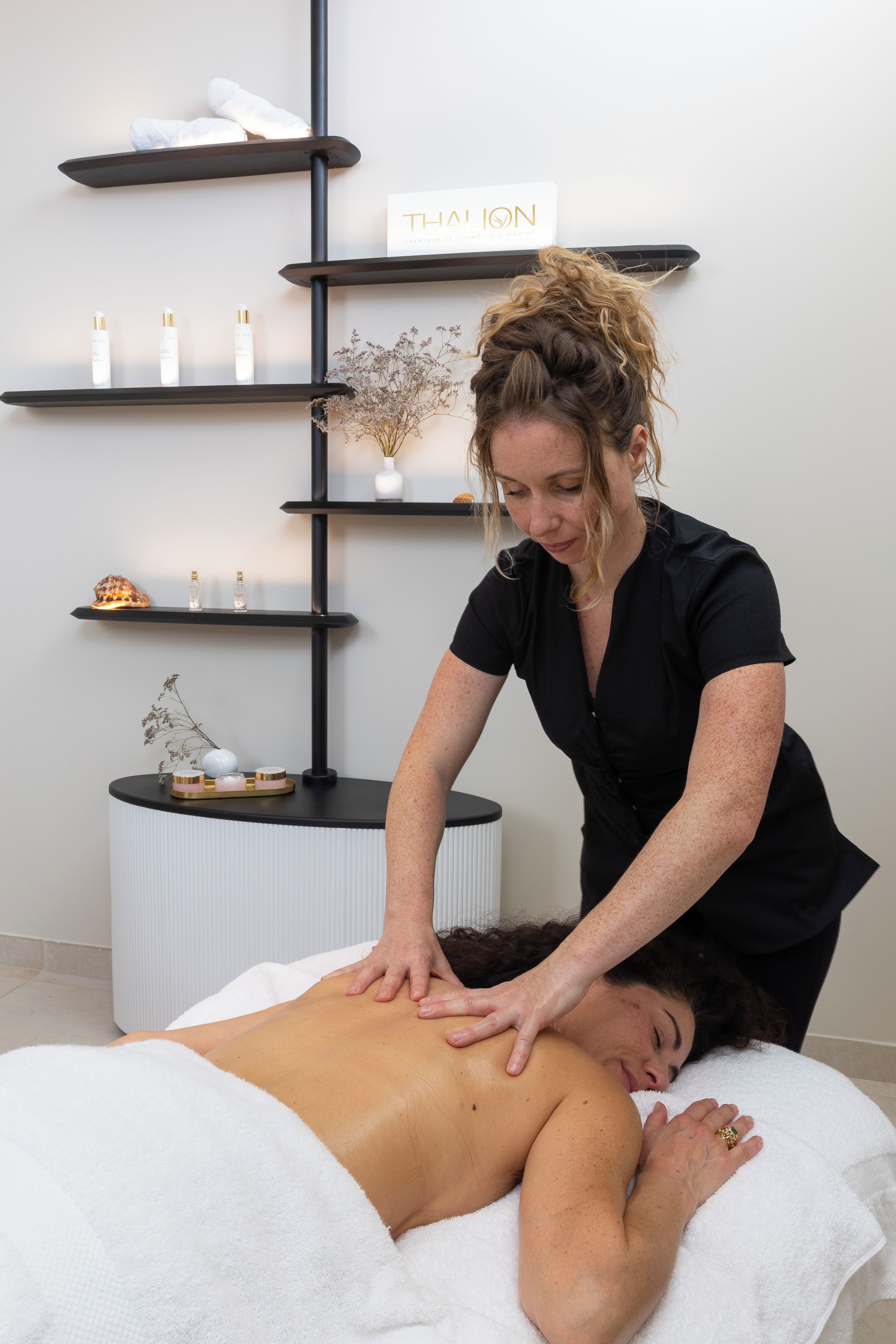 Eine Massageanwendung in einer Wellnessumgebung mit einem Regal, das verschiedene Produkte präsentiert, und einem Schild mit der Aufschrift 'THALION'.