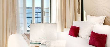 A hotel room with a bed, a window with a view of the sea, a towel, slippers, and a brochure.