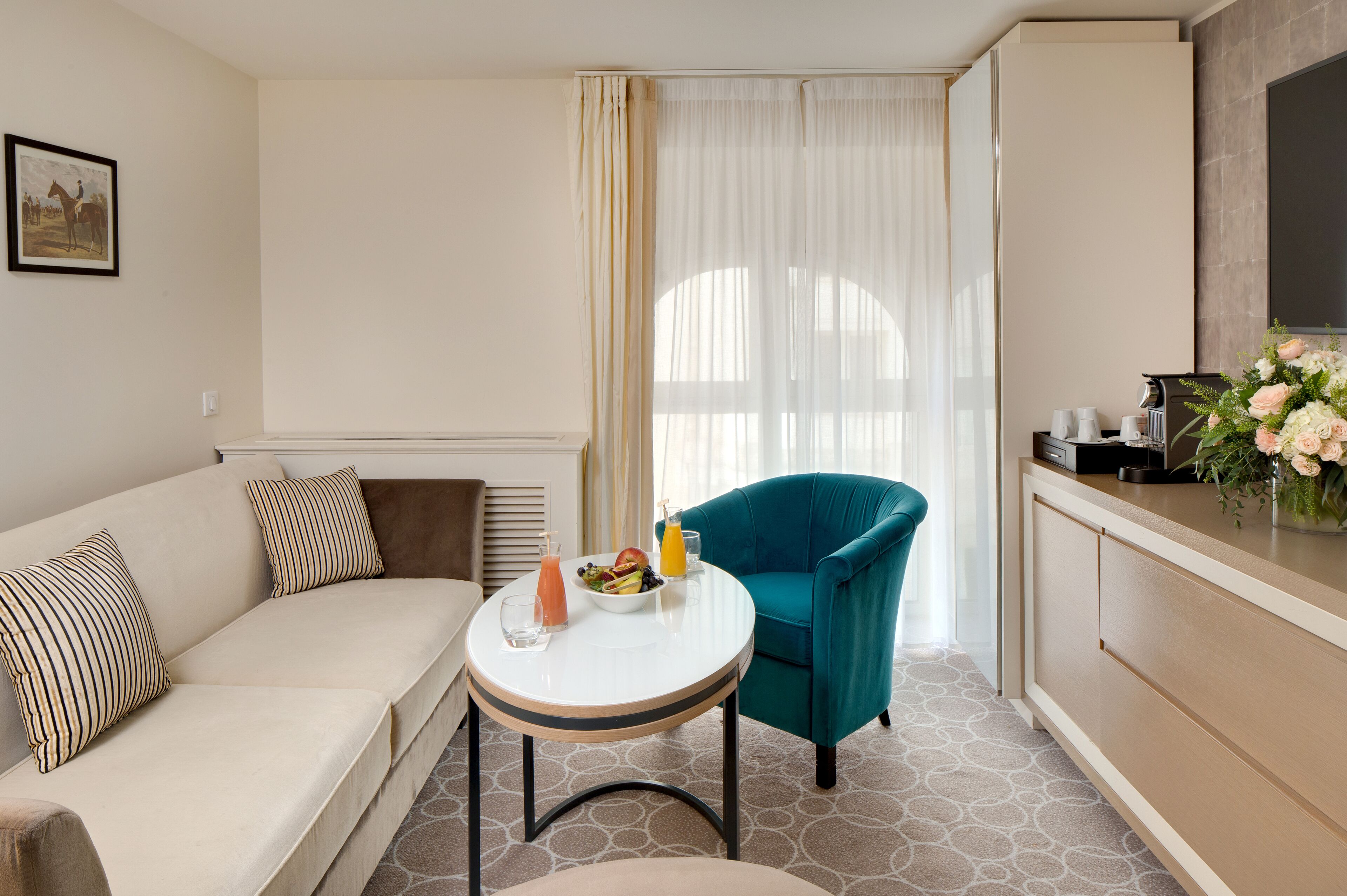 A modern hotel room with a sofa, a teal armchair, a small round table with a drink and fruit, and a kitchenette with a TV and flowers.