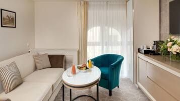 A modern hotel room with a sofa, a teal armchair, a small round table with a drink and fruit, and a kitchenette with a TV and flowers.