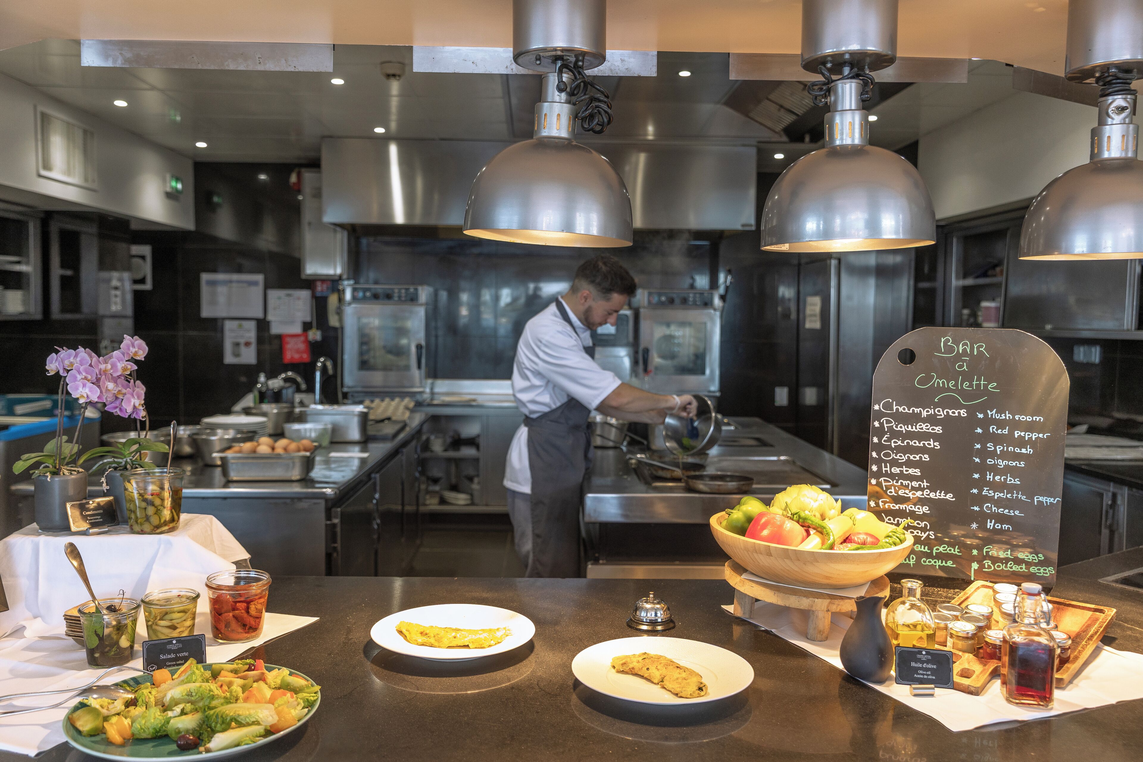 Un espace cuisine où un chef prépare des plats, un tableau affichant le menu du petit-déjeuner, et une présentation de fruits et légumes frais.