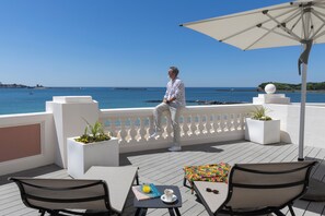 A man sitting on a balcony with a view of the sea, a white umbrella, and a table with a book and a glass of juice.