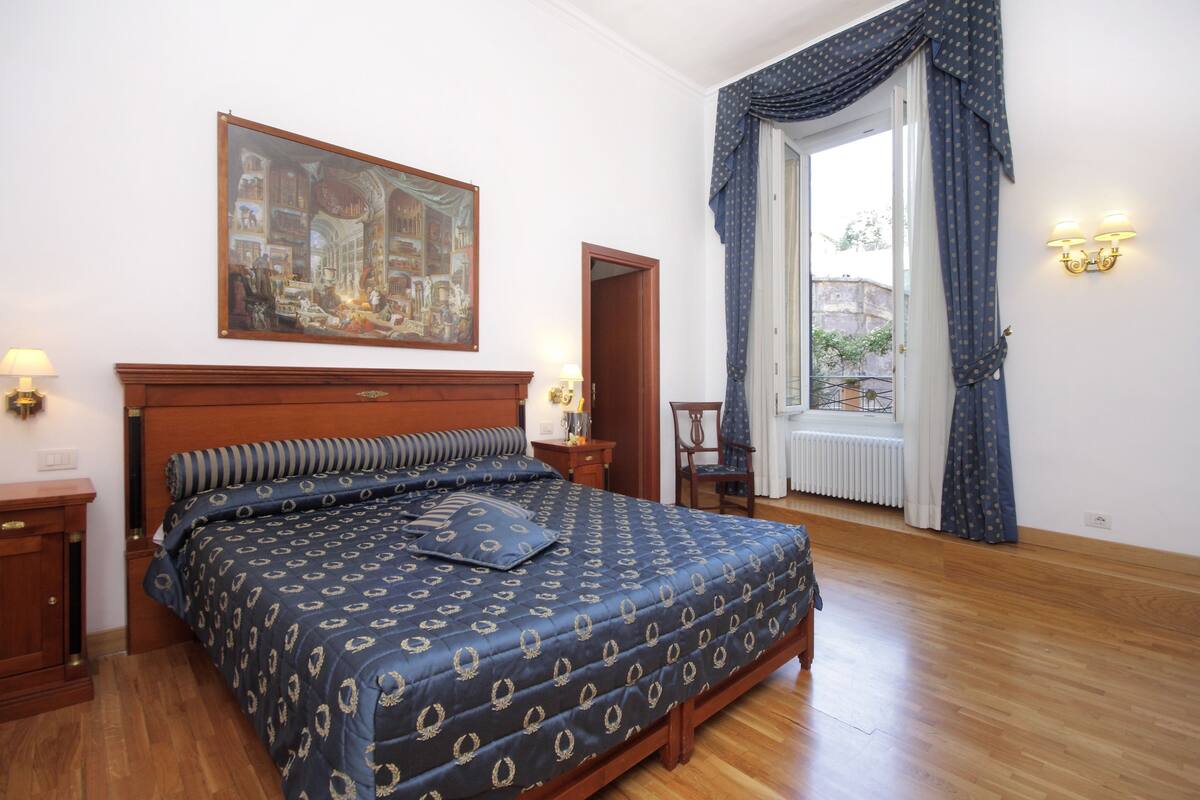 A bedroom with a large bed, a wooden headboard, a painting above the bed, a nightstand, a chair, and a window with curtains.