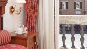A hotel room with a bed, a wooden nightstand with a cup and saucer, a red patterned curtain, and a view of a street with a sign reading "Piazza". - Relais Pierret (Rome)