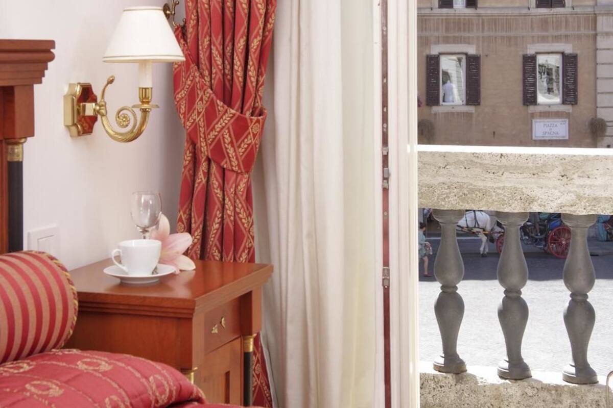 A hotel room with a bed, a wooden nightstand with a cup and saucer, a red patterned curtain, and a view of a street with a sign reading "Piazza".