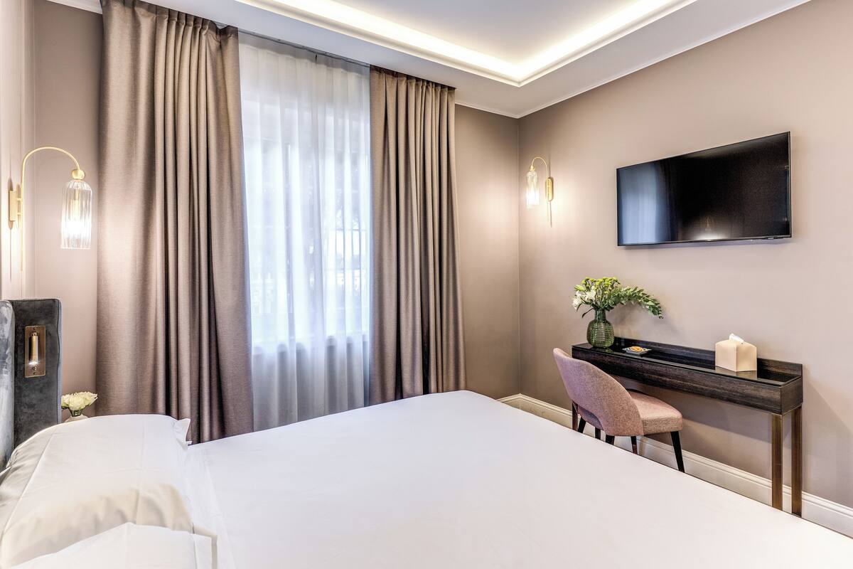 A modern hotel room with a large bed, a desk with a chair, a flat-screen TV, and a large window with curtains.