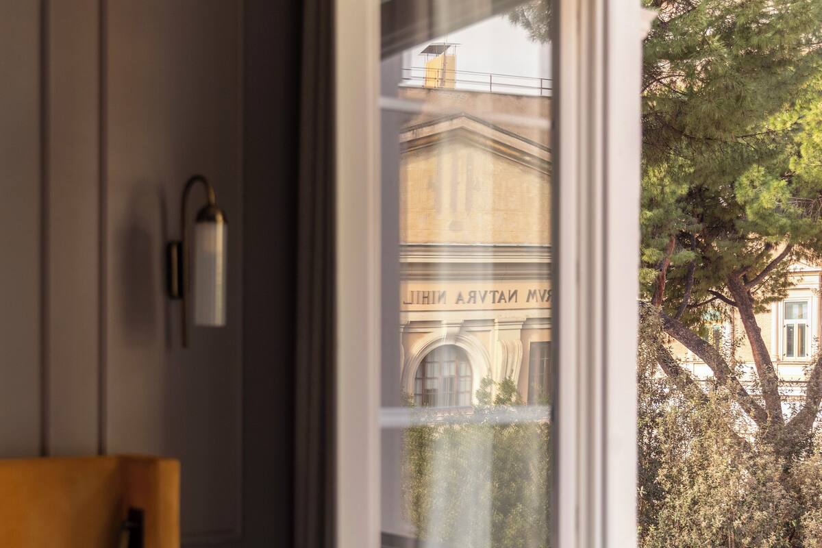 A hotel room with a bed, bedside table, lamp, and a window with a view of a building and trees.