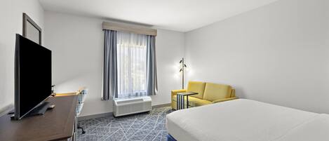 A hotel room with a bed, a TV, a desk, a chair, a lamp, and a window with curtains.