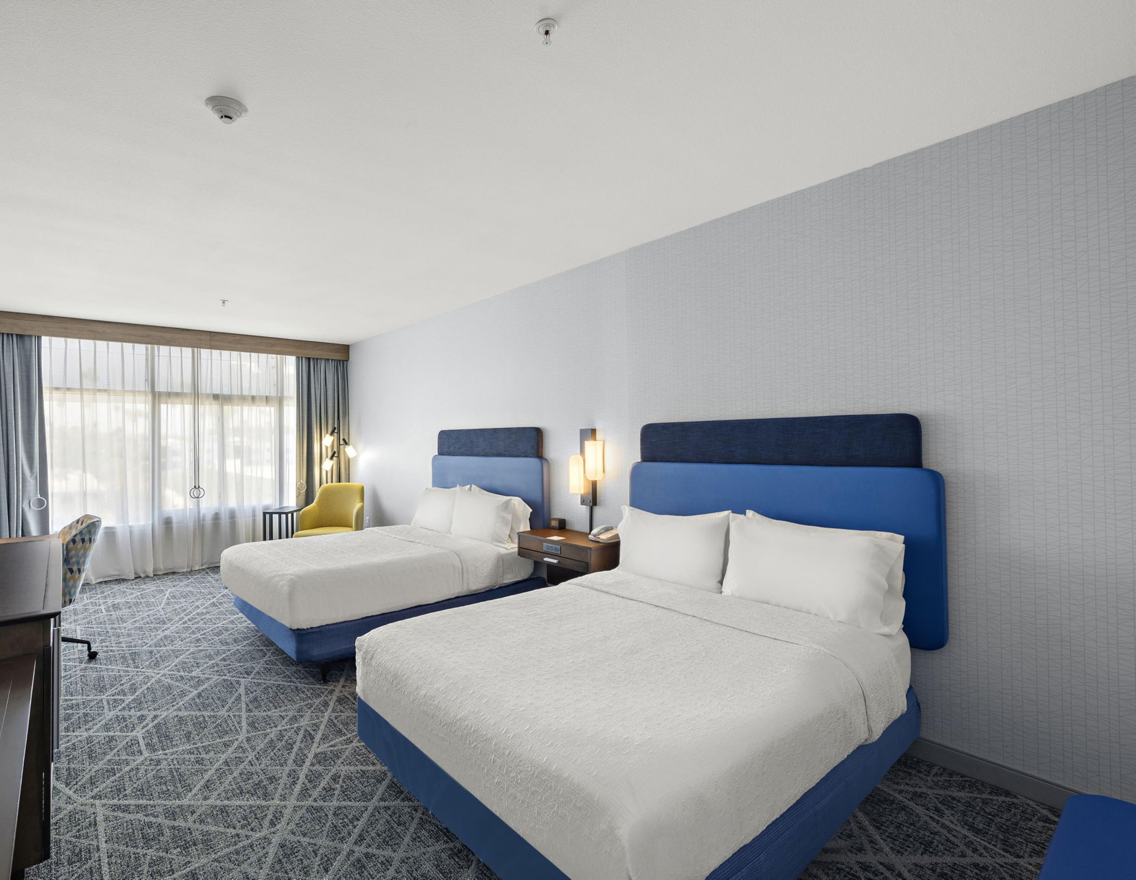 A hotel room with two beds, a desk, a chair, and a window with curtains.