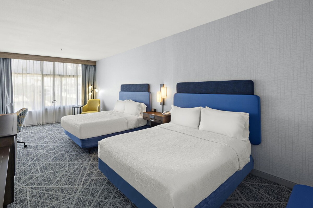 A hotel room with two beds, a desk, a chair, and a window with curtains.