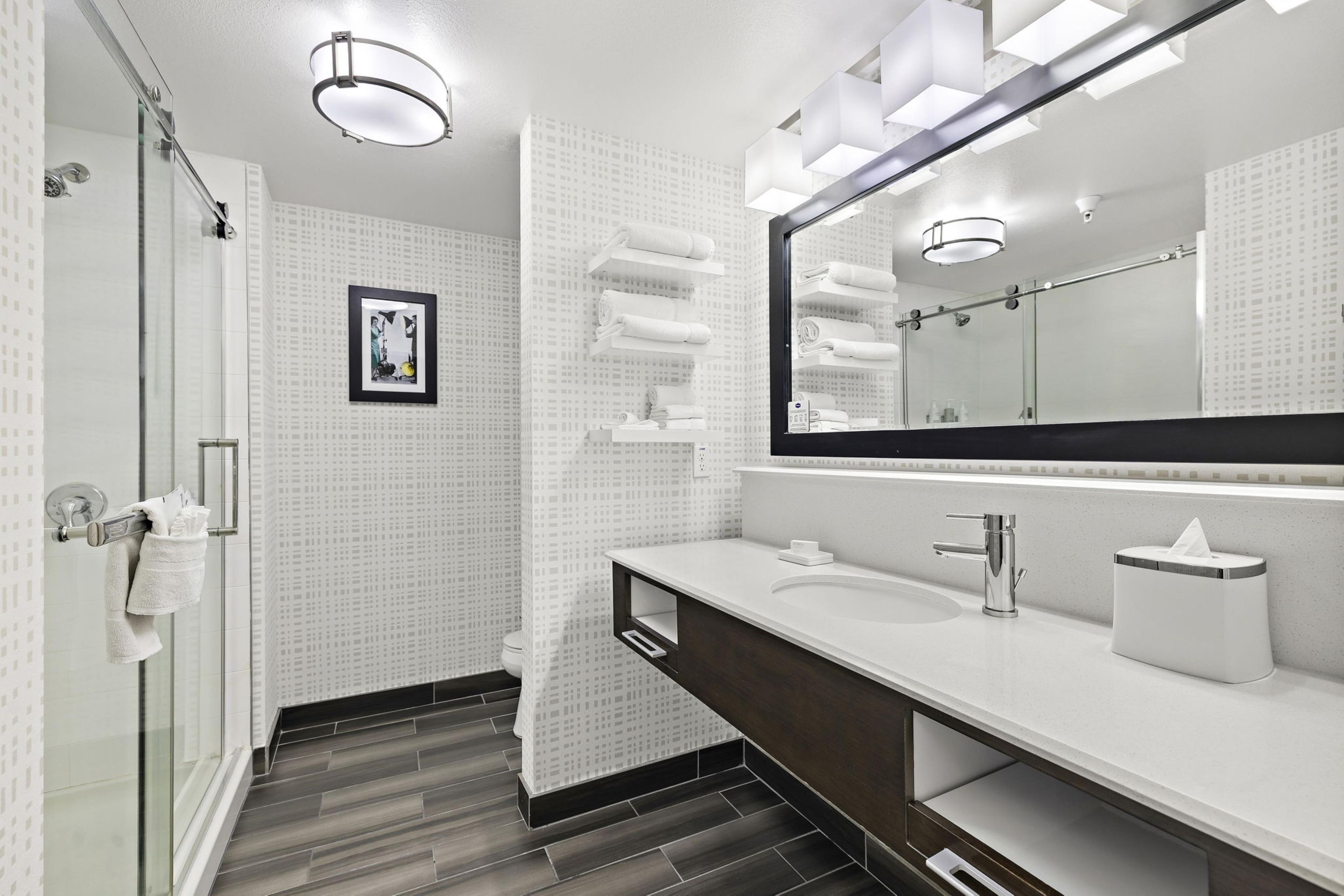 Un baño moderno con mampara de vidrio, un espejo grande y un tocador blanco con un mueble oscuro.