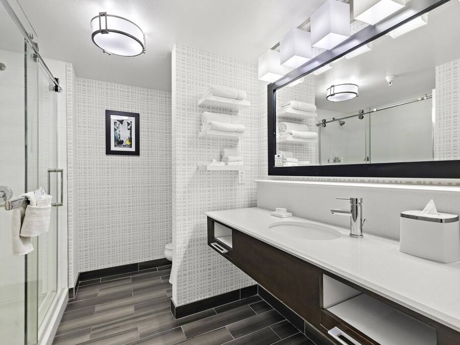 Un baño moderno con mampara de vidrio, un espejo grande y un tocador blanco con un mueble oscuro.
