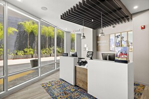 Zona de recepción de un hotel con diseño moderno, que incluye un mostrador de recepción, un cartel con la leyenda 'Welcome to Hampton' y vistas al exterior a través de amplios ventanales.