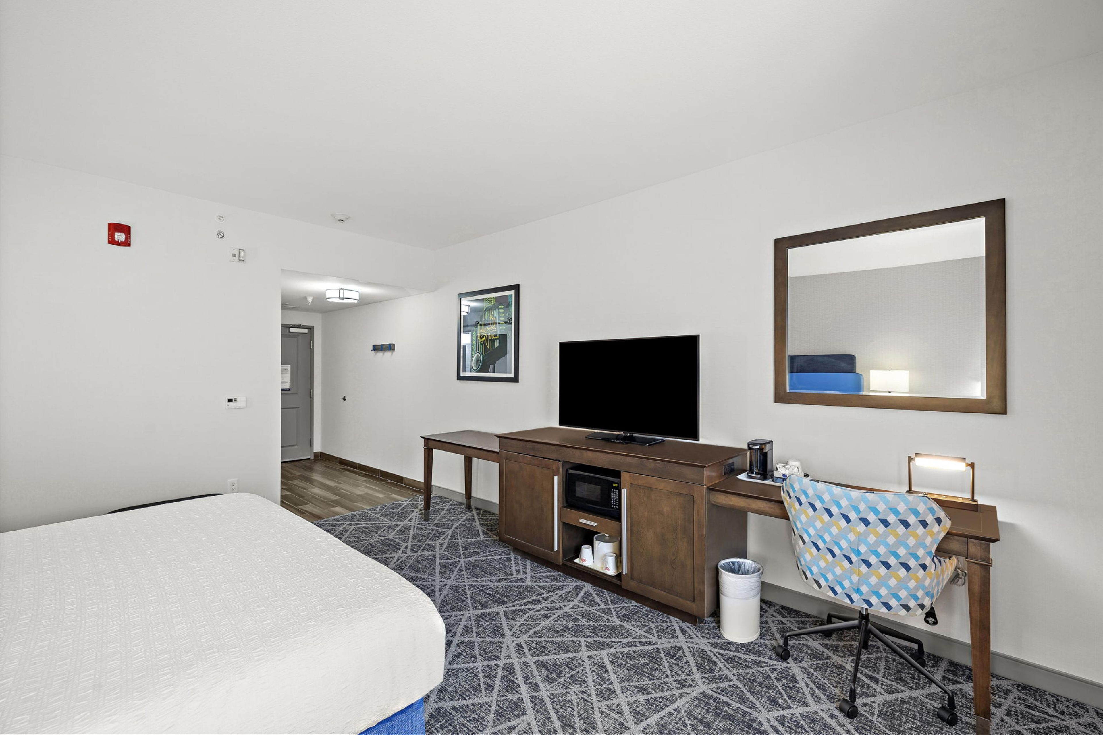Habitación de hotel con cama, escritorio con televisor de pantalla plana, una silla, un cesto de basura y un espejo en la pared.