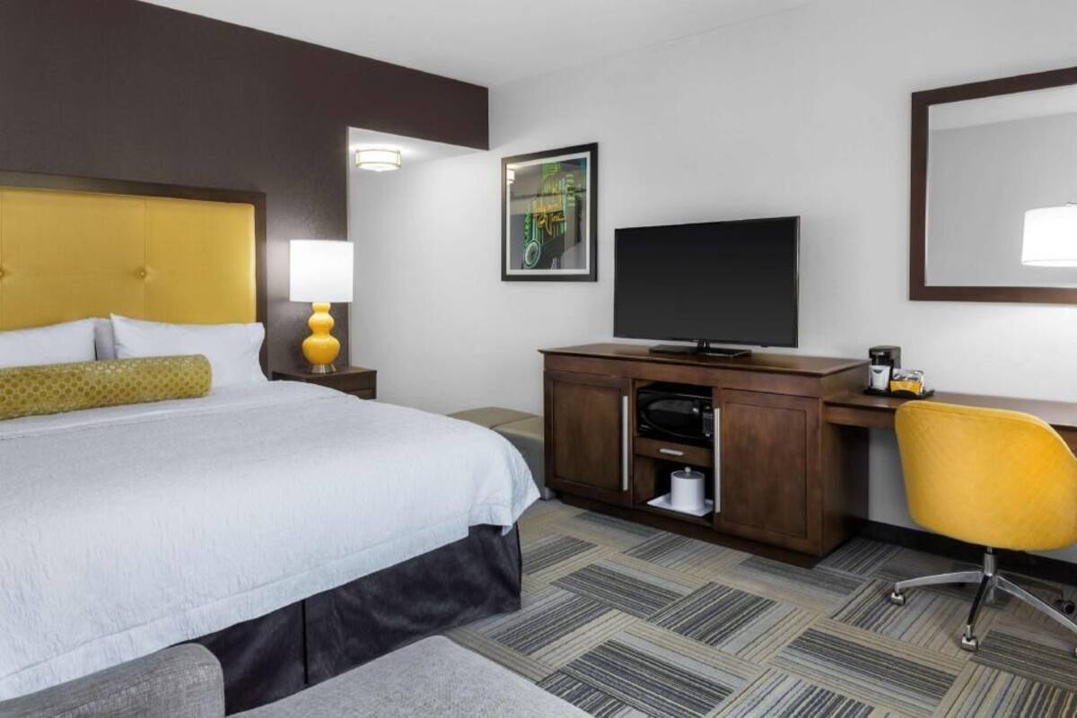 A hotel room with a bed, a desk, a chair, a TV, a lamp, a mirror, and a framed picture on the wall.
