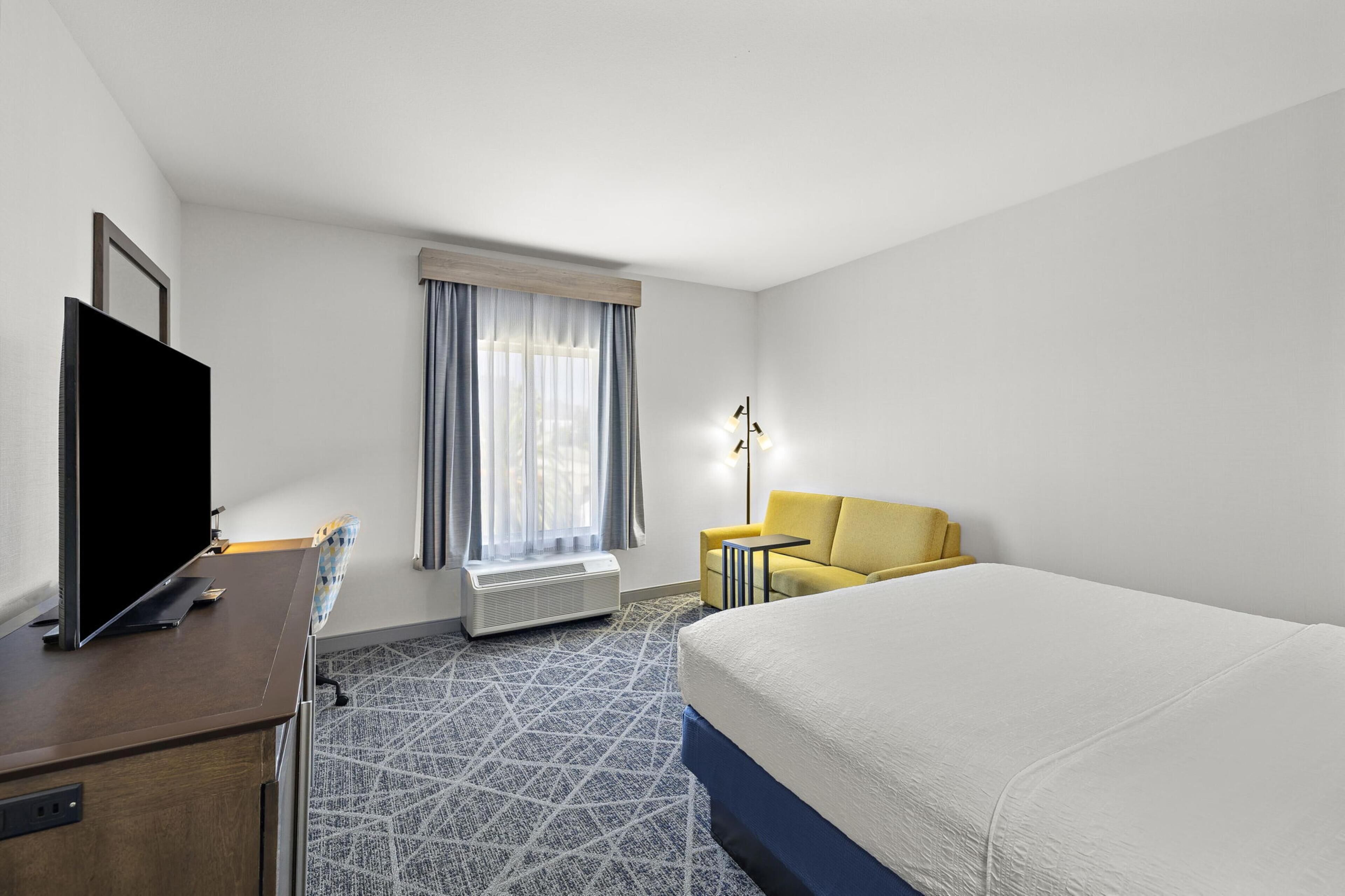 Een hotelkamer met een bed, televisie, bureau, stoel, lamp en een raam met gordijnen.
