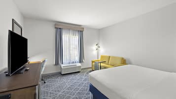 Quarto de hotel com cama, televisão, escrivaninha, cadeira, luminária e janela com cortinas.