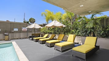 Zona junto a la piscina con sillones de descanso y sombrillas, cielo despejado y un letrero de advertencia en una cerca.