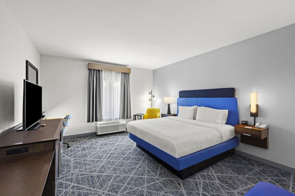 A hotel room with a bed, a desk, a television, a window with curtains, and a lamp.
