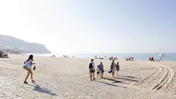 Une plage de sable où l’on voit des gens se promener et se tenir debout, un ciel dégagé et des bateaux sur l’eau.
