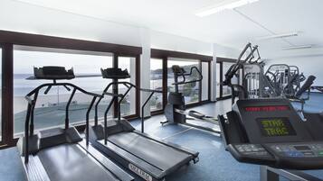 A gym with treadmills, ellipticals, and weight machines, featuring large windows with a view of the beach.